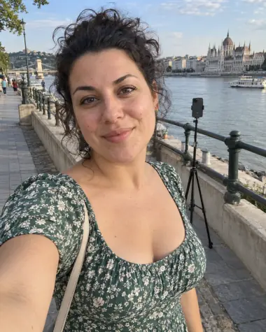 MI-illusztráció: kitalált felnőtt nő nyári ruhában készít szelfit a budapesti Duna-parton, mögötte telefonállvány látható.