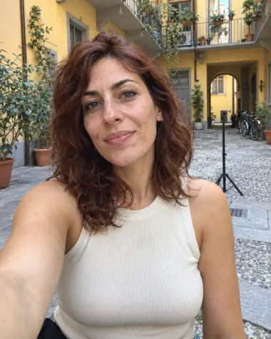 Illustrazione IA: una donna adulta immaginaria con capelli ramati scatta un selfie in un cortile milanese, con un treppiede alle spalle.