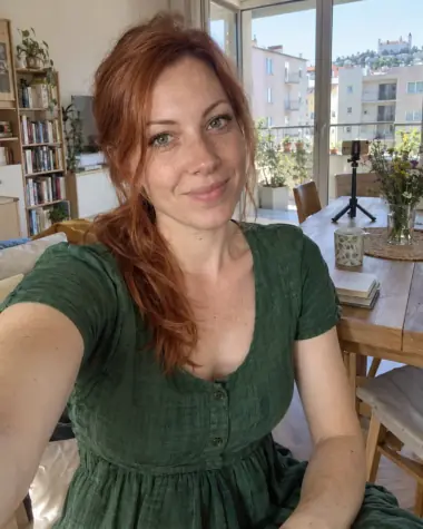 Ilustračná selfie fiktívnej dospelej ženy s ryšavými vlasmi a zelenými šatami v bratislavskom byte; v pozadí je statív.